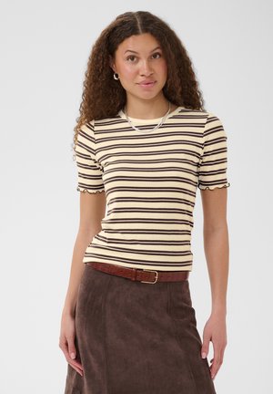 Jeune femme aux cheveux bouclés portant un t-shirt à manches courtes rayé crème et marron, une jupe marron et un collier en argent sur un fond uni.