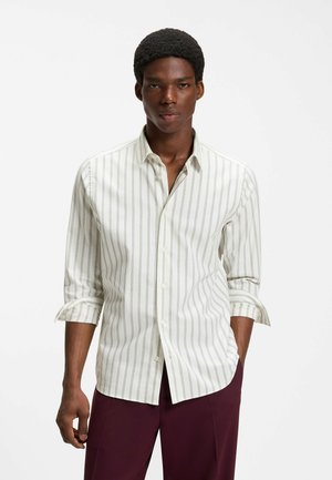 Man som bär en vit långärmad skjorta med tunna vertikala ränder och burgundyfärgade byxor, stående med ena handen i fickan.