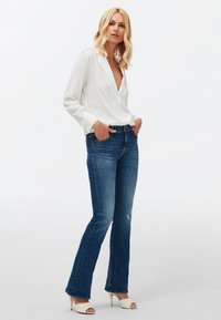 Chemisier blanc à manches longues avec un décolleté en V profond, associé à un jean évasé bleu légèrement déchiré, et des talons ouverts blancs.