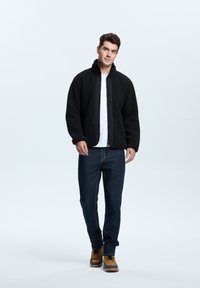 Musta fleece-takki, jossa on korkea kaulus ja vetoketju, yhdistettynä sinisiin farkkuihin ja ruskeisiin nauhakenkään. Pinnalla on sileä rakenne.