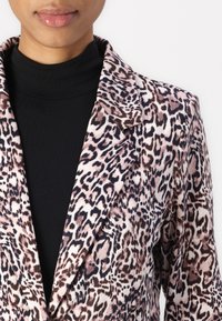 Blazer s leopardím vzorem v růžových a černých odstínech, vyrobený z hladké látky. Má šálový límec a dlouhé rukávy přes černý rolák.