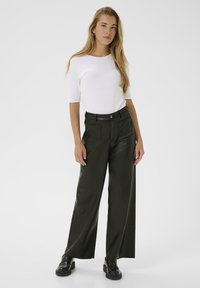 Ung kvinde står iført en hvid kortærmet bluse, sorte vide læderbukser, sorte klodsede loafers og sølvfarvede hoops.