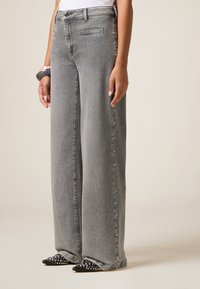 Vaqueros gris de pierna ancha con dos bolsillos frontales, detalles de cierre a los lados y una textura de denim suave. Combinados con zapatos negros de punta adornados con tachuelas.