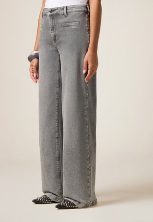 Graue Schlagjeans mit zwei Fronttaschen, seitlichen Druckknopfdetails und einer weichen Denim-Textur. Kombiniert mit spitzen schwarzen Schuhen, die mit Nieten verziert sind.