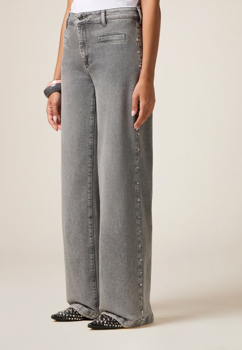 Vaqueros gris de pierna ancha con dos bolsillos frontales, detalles de cierre a los lados y una textura de denim suave. Combinados con zapatos negros de punta adornados con tachuelas.