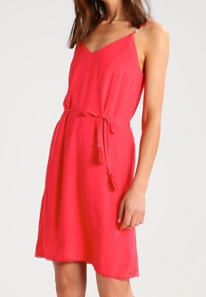 Mujer con un vestido rojo sin mangas hasta la rodilla, con un cinturón fino atado a la cintura y un delicado adorno en el dobladillo.