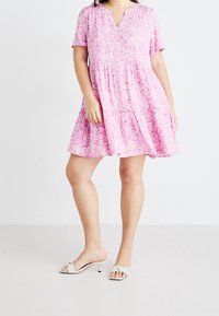 Růžové květinové šaty s výstřihem do V, krátkými rukávy a vrstvenou sukní. Hladká látka s lehkou texturou. Kombinované s béžovými nazouvacími sandály.