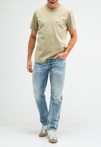 Licht olijfgroene t-shirt met korte mouwen en een ronde halslijn, voorzien van een klein bedrukt logo op de linkerkant van de borst, gecombineerd met vervaagde blauwe jeans en grijze sneakers.