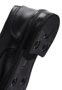 Chaussure en cuir noir avec un bout arrondi, semelle robuste dotée d'accents en caoutchouc circulaires et d'un detailing texturé le long du bord supérieur.