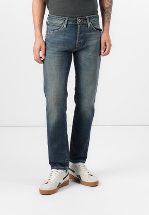 Man die versleten blauwe jeans en witte sneakers draagt, staand tegen een eenvoudige lichte achtergrond.