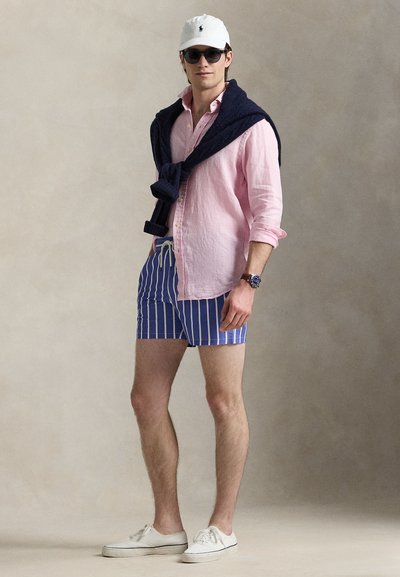 Trumpomis rankovėmis rožinės spalvos lininė marškiniai, ant viršaus užmesta tamsiai mėlyna megztinis, dryžuotos mėlynos maudymosi glaudės, balti sportbačiai, balta kepurė ir akiniai nuo saulės. Kasdienis vasaros įvaizdis.