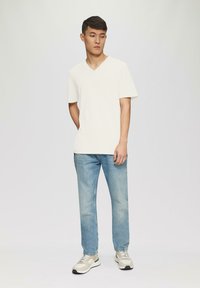 Weißes V-Ausschnitt-T-Shirt, kurze Ärmel, kombiniert mit hellblauen Jeans und beigen Sneakern, vor einem schlichten hellen Hintergrund.