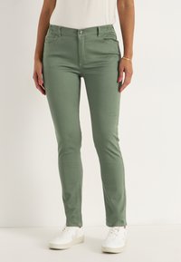 Vihreät puuvillasekoitehousut, joissa on istuva muotoilu, etutaskut ja nappikiinnitys vyötäröllä, yhdistettynä valkoisiin lenkkareihin.