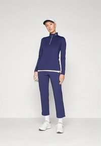 Σετ αθλητικό σε ναυτικό μπλε χρώμα με μακρυμάνικο μπλουζάκι με φερμουάρ και cropped παντελόνια, κατασκευασμένο από ελαστικό ύφασμα, συνδυασμένο με λευκά αθλητικά παπούτσια και καπέλο.