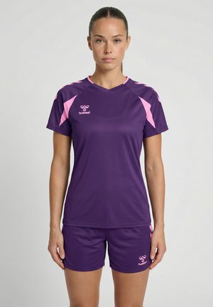 Femme portant un maillot de sport violet et un short avec des touches roses, debout face à un fond uni.