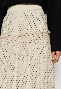 Jupe beige à pois noirs, taille élastique et couture à volants, portée avec un haut à manches longues noir.