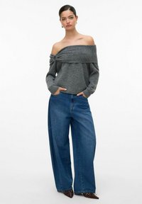 Grauer schulterfreier Pullover mit einem verdrehten Detail, kombiniert mit weiten blauen Jeans. Das Outfit hat lange Ärmel und einen lässigen Schnitt.