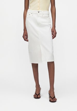 Žena v bielej džínsovej sukni po kolená, béžovom tielku bez rukávov a čiernych sandáloch na opätku stojí pred jednofarebným pozadím.