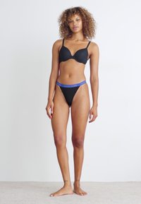 Zwart lingeriesetje met een gewatteerde, onderbedekte bh en een string. De bh heeft dunne bandjes; de string heeft een blauwe merkband. Soepele stoftextuur.