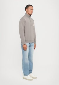 Sudadera gris de cremallera con cuello alto, combinada con jeans rectos de color azul claro y zapatillas blancas. Diseño práctico y casual.