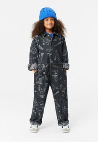 Jeansoverall i mörkgrått med grafiska mönster, långa ärmar uppvikta och dragkedja fram; bärs med en blå stickad mössa och vita sneakers.