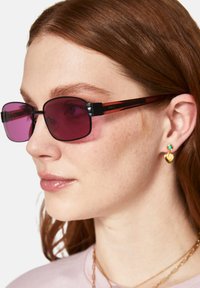 Lila tonade rektangulära solglasögon med svarta och röda ramlister. Bärs av en person som visar upp små örhängen i guld i form av hjärtan och turkosa.