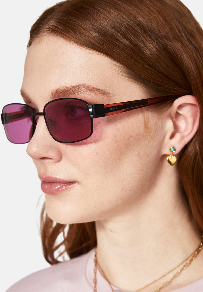 Lila tonade rektangulära solglasögon med svarta och röda ramlister. Bärs av en person som visar upp små örhängen i guld i form av hjärtan och turkosa.