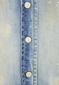 Camicia di jeans azzurro chiaro con bottoni bianchi, caratterizzata da cuciture a contrasto lungo il collo e una superficie sbiadita e testurizzata con macchie.