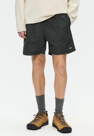 Osoba koja nosi crne kratke hlače, bež košulju s dugim rukavima, sive čarape i smeđe planinarske cipele stoji na bijeloj pozadini.