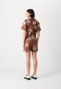 Jumpsuit corto marrone con maniche corte, caratterizzato da motivi astratti bianchi e una vita cintrata, dalla vestibilità rilassata e texture liscia.