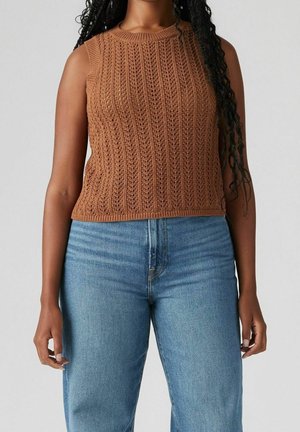 Femme portant un haut en tricot marron sans manches et un jean bleu clair taille haute, debout devant un fond gris uni.