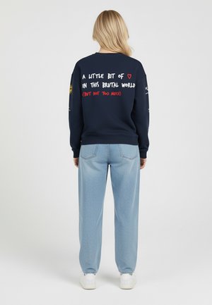 Felpa blu navy con testo bianco e rosso sulla schiena, abbinata a pantaloni leggeri blu chiaro con fantasia. L'outfit presenta una vestibilità rilassata e casual.
