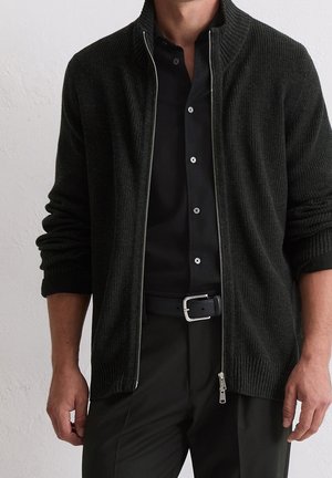 Uomo con camicia nera abbottonata, cardigan nero lavorato a maglia con zip e maniche arrotolate, cintura nera e pantaloni neri davanti a una parete bianca.
