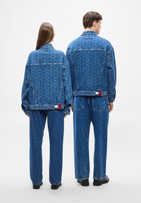 Traper jakna i široke traperice od plavog materijala, s ponovljenim uzorkom teksta i kontrastnim oznakama brenda u crvenoj, bijeloj i plavoj boji.