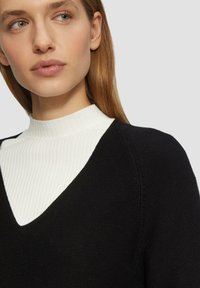 Zwarte gebreide trui met een V-hals, laag over een witte geribbelde coltrui, waarbij contrasterende texturen en strakke lijnen worden getoond.