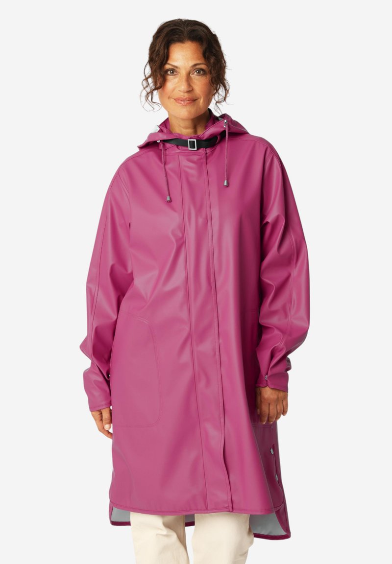 Femme portant un long imperméable rose ample avec une capuche et des cordons réglables, debout devant un fond uni.