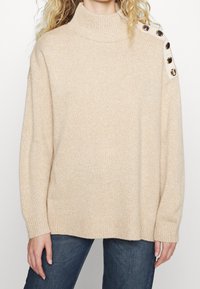 Beżowy sweter z golfem wykonany z dzianiny, o luźnym kroju, z guziczkami na ramieniu oraz prążkowanymi mankietami i dołem.