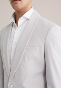 Heledad hall hall blazer sileva tekstuuriga, struktureeritud kraega ja ühe taskuga; kantud valge sviiteri üle, millel on nähtavad nööbid.