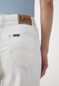 Jean en denim blanc avec une pièce en cuir marquée "Lee" au niveau de la taille arrière, une petite étiquette sur la poche arrière et des détails de couture subtils.