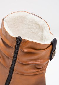 Bottine en cuir marron avec un haut arrondi, doublure douce blanche, et un zip noir vertical sur le côté, avec un détail de bouton décoratif.