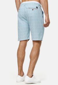 Lichtblauwe geruite shorts van zachte stof, met een knoopzak aan de achterkant en zijzakken. Draagt witte sneakers.