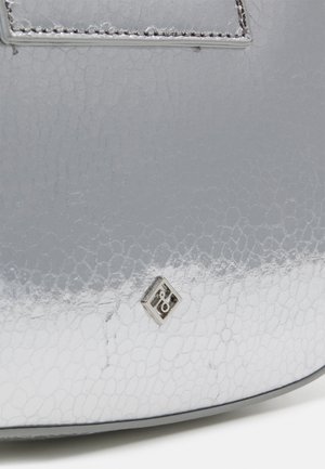 Nahaufnahme der strukturierten Oberfläche einer silbernen Handtasche mit kleinem quadratischem Metall-Logo und sichtbarer Naht am Rand.