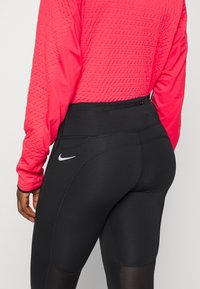Gros plan sur une personne portant des leggings noirs Nike avec une poche zippée sur le côté et un haut rouge à manches longues texturé.
