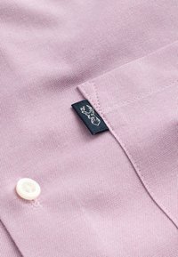 Material de cămașă țesut în nuanță de violet deschis, cu textură netedă. Prezintă un nasture alb și o etichetă neagră cu un logo cu cerb pe buzunar.
