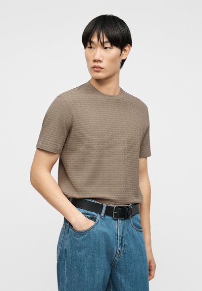 Νεαρός άνδρας με μαύρα μαλλιά, φορώντας καφέ πουκάμισο με κοντό μανίκι και υφή, μπαίνει μέσα σε μπλε τζιν με μαύρη ζώνη, στέκεται ενάντια σε μια απλή φόντο.