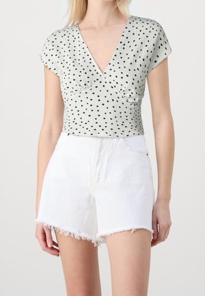 Mujer con una blusa blanca de cuello en V con lunares negros y shorts de mezclilla blancos de tiro alto con dobladillos deshilachados, posando contra un fondo liso.