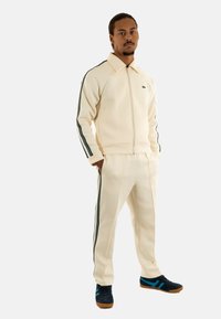 Chándal deportivo en crema con rayas verde oscuro, que incluye una chaqueta con cremallera y pantalones a juego, fabricado con tejido suave, acompañado de zapatillas negras.