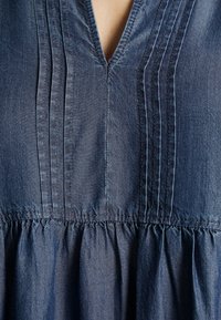 Denim-toppi, jossa on V-pääntie, pystysuorat pleatit ja kerätty vyötärö. Kangas on teksturoitu tummansinisellä sävyllä.
