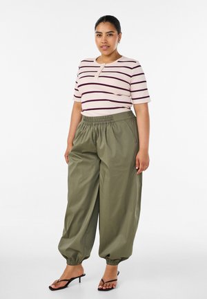 Vrouw staat, draagt een beige-paars gestreepte top met korte mouwen, olijfgroene wijd uitlopende broek met elastische taille en zwarte open teen sandalen met hak.