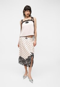 Femme portant un caraco en satin blanc avec dentelle noire et une jupe midi blanche à pois noirs, bordée de dentelle, fendue sur le côté, et des chaussures argentées à bouts pointus.